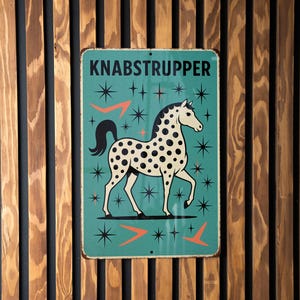 May include: A vintage-style metal sign with the word "KNABSTRUPPER" at the top. The sign features a white horse with black spots, surrounded by starbursts and orange arrow designs, set against a teal background. The sign is mounted on a wooden slatted wall.