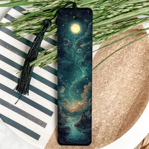 May include: A dark blue bookmark with a colorful galaxy scene and a single person standing on a cliff overlooking a river. The bookmark has a black tassel.