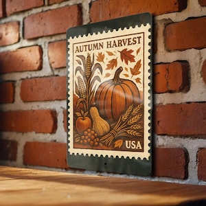 May include: A rectangular metal sign with a vintage design featuring the words "AUTUMN HARVEST" and "USA". The artwork includes a pumpkin, corn, leaves, an apple, and a gourd in shades of orange, brown, and beige. The sign is displayed against a brick wall.