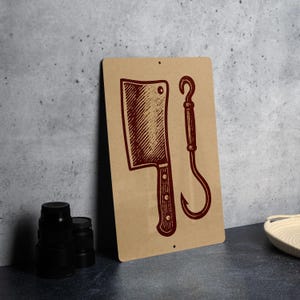 May include: A rectangular metal sign with a butcher theme. The sign features a cleaver and a meat hook in a dark red color against a tan background. The sign is mounted on a gray wall.