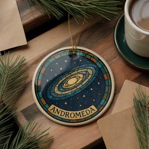 May include: Round ceramic ornament featuring a galaxy design with the word "ANDROMEDA" in a banner. The ornament has a gold string for hanging and is decorated with blue, gold, and teal colors. The ornament is on a wooden surface.