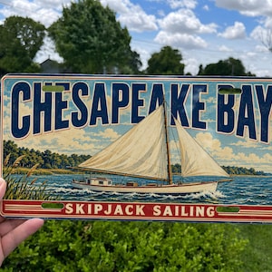 May include: A rectangular metal sign featuring a vintage-style illustration of a white sailboat on blue water. The text CHESAPEAKE BAY is printed in large blue letters at the top, with the words SKIPJACK SAILING in red at the bottom of the sign.