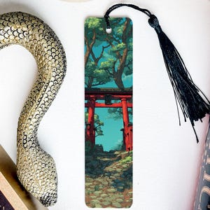 May include: A bookmark featuring a Japanese torii gate scene with a black tassel. A decorative golden snake and a book with the word "PRACTIC" are also visible. The bookmark has a detailed illustration of a path leading to the gate.