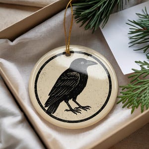May include: Round ceramic ornament featuring a black crow illustration on a cream background, framed by a black border. The ornament has a gold string for hanging and is presented in a gift box.