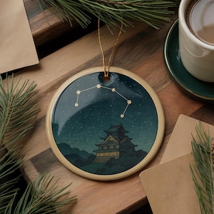 May include: Round ceramic ornament featuring a night scene with a castle and constellation design. The ornament has a gold string for hanging and a cream-colored border. The background is dark blue with white stars.