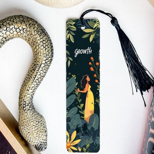 May include: A decorative bookmark with a black tassel. The bookmark features a woman in a yellow dress surrounded by foliage, with the word "Growth" at the top. The background is dark green with orange and yellow flowers.