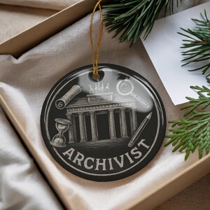 May include: Black round ornament with a gold string, featuring a detailed white illustration of a building, hourglass, magnifying glass, and pencil. The word "ARCHIVIST" is written in white. The ornament is in a gift box.