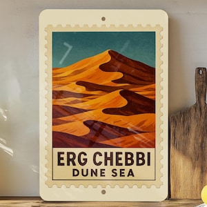 May include: A rectangular wall art piece featuring a desert landscape with orange sand dunes under a teal sky. The text "ERG CHEBBI DUNE SEA" is displayed in a rectangular frame at the bottom. The art piece has a postage stamp design.