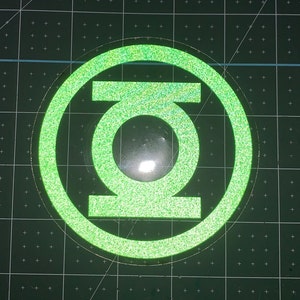 May include: Green and white glitter Green Lantern logo sticker. The logo is a circle with a stylized letter G inside.