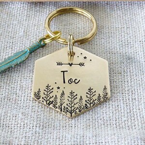 Op de afbeelding: Een goudkleurige, zeshoekige sleutelhanger met de naam "Toc" gegraveerd, met een bosontwerp en een turquoise veerketting. De sleutelhanger is bevestigd aan een sleutelring en rust op een linnen zakje. Het label luidt "WOLF & BEAR HANDMADE IN AUSTRIA."