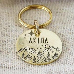 May include: A round gold-colored metal pet tag with a split ring, featuring an engraved mountain and forest landscape, a sun, stars, and the name AKIMA. The tag is placed on a small beige fabric pouch with a brown paper tag labeled WOLF & BEAR.