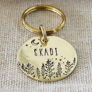 Puede incluir: Una placa redonda de latón para mascotas con una anilla, que presenta el nombre SKADI grabado, una luna creciente, estrellas y un bosque de pinos. La placa se presenta con una etiqueta de papel marrón WOLF & BEAR HANDMADE IN AUSTRIA y una bolsa.