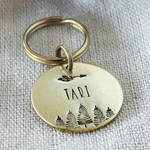 Op de afbeelding: Een ronde, messingkleurige sleutelhanger met de naam "Tari" en een bosontwerp. De tag is bevestigd aan een sleutelring en rust op een klein, beige stoffen zakje. De tag is gestempeld met de woorden "WOLF & BEAR HANDMADE IN AUSTRIA" op een bruin label.