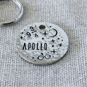 May include: A round, silver-coloured metal pet ID tag with a hammered texture, featuring the name APOLLO, a 9 3/4 symbol, a crescent moon, stars, and glasses. A silver split ring is included, and the tag is presented with a WOLF & BEAR brand tag.