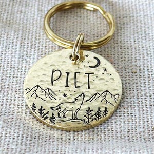 Könnte beinhalten: Eine runde Haustiermarke aus Messing mit gehämmertem Finish, graviert mit einem Wolf, Bergen, Bäumen und einer Mondsichel. Der Name PIET ist mittig eingeprägt. Die Marke verfügt über einen goldenen Metallring zur Befestigung am Halsband.