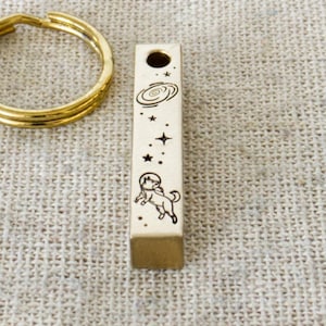 May include: A gold key ring and a rectangular pendant with an astronaut and celestial design are displayed on a linen pouch. The pendant has a hole at the top and is engraved with stars, planets, and an astronaut. A brown tag with the text "WOLF & BEAR HANDMADE IN AUSTRIA" is in the background.
