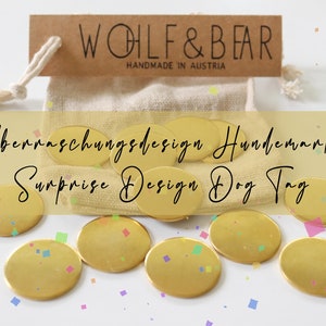 Puede incluir: Se muestran chapas doradas para perros, con el texto "Surprise Design Dog Tag" y "WOLF & BEAR HANDMADE IN AUSTRIA". Las chapas están frente a una pequeña bolsa beige y una etiqueta marrón.