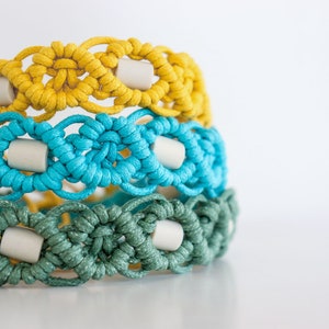 May include: Three macrame bracelets stacked on top of each other. The top bracelet is yellow, the middle is turquoise, and the bottom is green. Each bracelet has a repeating pattern with off-white cylindrical beads. The bracelets are made of knotted cord.