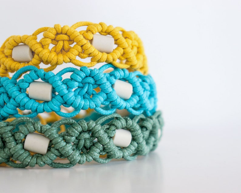 May include: Three macrame bracelets stacked on top of each other. The top bracelet is yellow, the middle is turquoise, and the bottom is green. Each bracelet has a repeating pattern with off-white cylindrical beads. The bracelets are made of knotted cord.