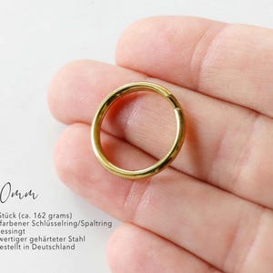 Może przedstawiać: Złoty metalowy pierścień typu split ring o średnicy 20mm. Tekst na obrazie wskazuje zestaw 100 sztuk o wadze około 162 gramów, wykonany z mosiądzowanej hartowanej stali i wyprodukowany w Niemczech.
