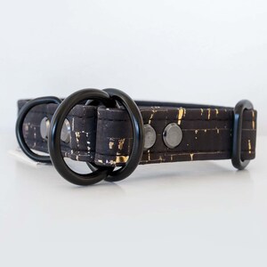 May include: Black dog collar with gold flecks and black hardware. The collar features two black metal D-rings and a black metal buckle. The collar is made of a textured material with a pattern of gold flecks.