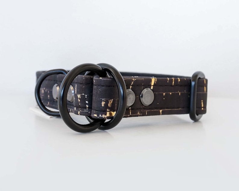 May include: Black dog collar with gold flecks and black hardware. The collar features two black metal D-rings and a black metal buckle. The collar is made of a textured material with a pattern of gold flecks.