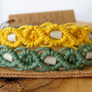 May include: Two macrame bracelets, one yellow and one green, are stacked. Each bracelet features a woven design with white cylindrical beads. The bracelets are displayed on a burlap surface.