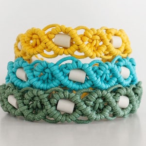 May include: Three macrame bracelets in yellow, turquoise, and green. Each bracelet features a woven design with a series of circular patterns and small, cylindrical beads. The bracelets are stacked, showcasing the different colors and textures.