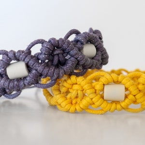 May include: Two macrame bracelets, one in a deep purple and the other in a bright yellow. Each bracelet features a repeating pattern with a light-colored cylindrical bead at the center of each design element. The bracelets are displayed on a white surface.