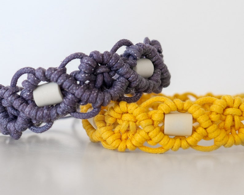 May include: Two macrame bracelets, one in a deep purple and the other in a bright yellow. Each bracelet features a repeating pattern with a light-colored cylindrical bead at the center of each design element. The bracelets are displayed on a white surface.