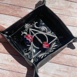 May include: Black dice tray with a silver glitter finish and an embroidered design of the Deathly Hallows symbol from the Harry Potter series. Two dice are inside the tray, one red and one black.