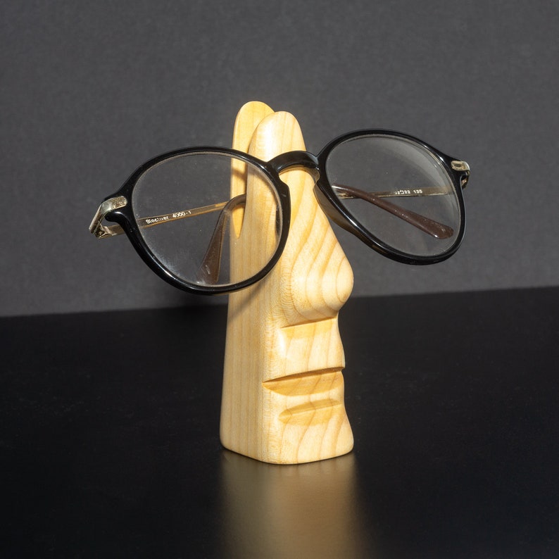 Puede incluir: Un soporte de madera con forma de cara con un par de gafas redondas negras apoyadas en la nariz.