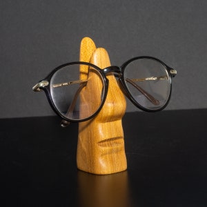 Puede incluir: Un soporte de madera con forma de cara con un par de gafas redondas negras apoyadas en &eacute;l.
