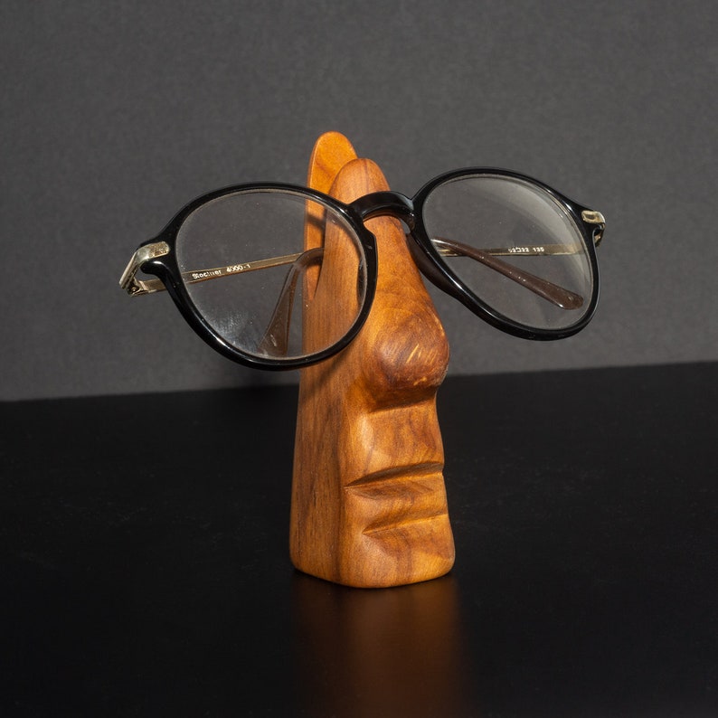 Puede incluir: Un soporte de madera con forma de cara con nariz y boca, que sostiene unas gafas con montura negra.