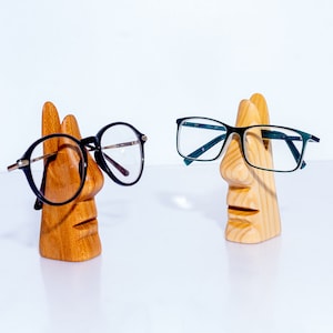 Puede incluir: Dos soportes para gafas de madera con forma de cara. Un soporte sostiene un par de gafas negras, y el otro sostiene un par de gafas verdes.