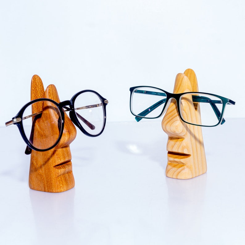 Puede incluir: Dos soportes para gafas de madera con forma de cara. Un soporte sostiene un par de gafas negras, y el otro sostiene un par de gafas verdes.