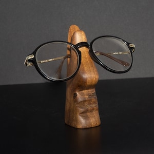 Puede incluir: Un soporte de madera con forma de cara con un par de gafas redondas negras apoyadas en la nariz.