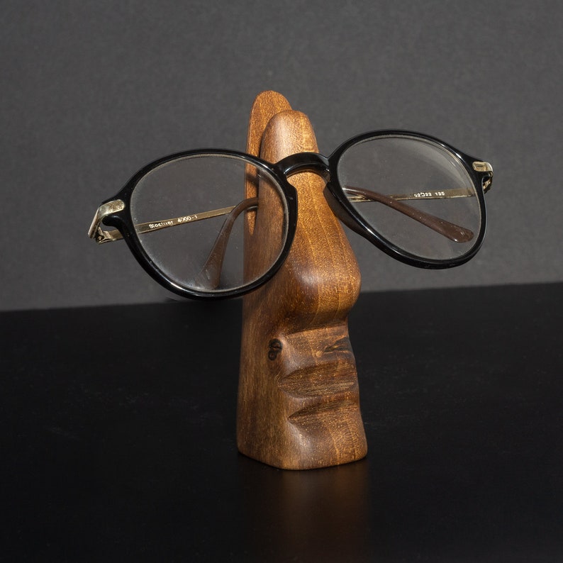 Puede incluir: Un soporte de madera con forma de cara con un par de gafas redondas negras apoyadas en la nariz.