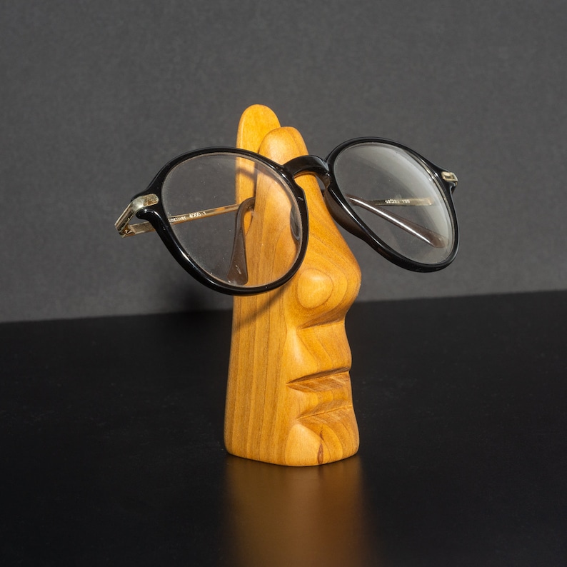 Puede incluir: Una escultura de madera de una cara con un par de gafas de montura negra apoyadas en la nariz. La escultura est&aacute; tallada con una cara detallada y tiene un acabado de madera natural.
