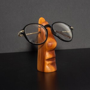 Puede incluir: Un soporte para gafas de madera con forma de cara, con nariz y boca talladas. El soporte sostiene un par de gafas con montura negra.
