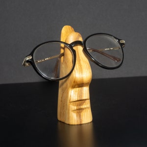 Puede incluir: Soporte de madera para gafas en forma de cara con boca y nariz talladas, sosteniendo un par de gafas redondas negras.