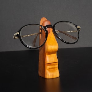 Puede incluir: Un soporte de madera con forma de cara con nariz y boca, que sostiene unas gafas con montura negra.