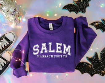Op de afbeelding: Een paarse sweatshirt met het woord "SALEM" in witte blokletters boven "MASSACHUSETTS". De sweatshirt is omgeven door Halloween-decoraties, waaronder zwarte vleermuisuitsparingen en een discobol schedel. Een paar zwart-witte sneakers staan ook op de foto.