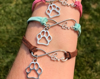 May include: Three infinity symbol bracelets with paw print charms and the word "love" on each. The bracelets are brown, pink, and turquoise.