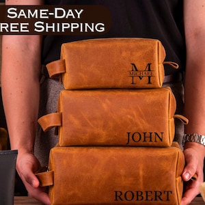 May include: Three brown leather toiletry bags with personalized names. The top bag has the name "Michael" with an initial "M" monogram. The middle bag has the name "John" and the bottom bag has the name "Robert".