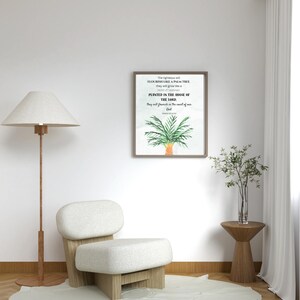 May include: A framed print with a palm tree illustration and text, set in a minimalist living room. The room includes a white chair, a wooden side table, a floor lamp, and a vase with greenery. The print's text reads: "The righteous will flourish like a palm tree..."