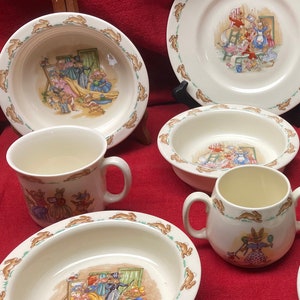 May include: A set of Beatrix Potter themed children's dishes, including a plate, bowl, and mug, featuring Peter Rabbit and other characters. The dishes are white with a colorful design.