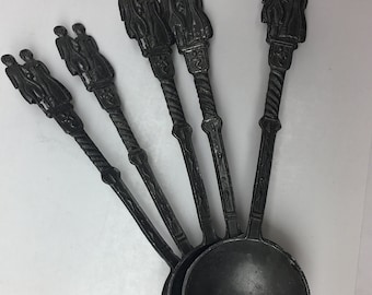 May include: Set of six pewter spoons with ornate handles featuring a pair of figures. The spoons are black and have a rounded bowl.
