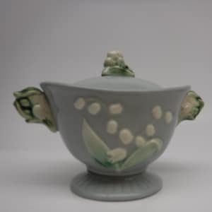 Può includere: Zuccheriera in ceramica azzurra con coperchio, con un motivo di mughetto. La ciotola ha una base a piedistallo e manici a forma di foglie. Il coperchio è sormontato da un grappolo di fiori di mughetto.