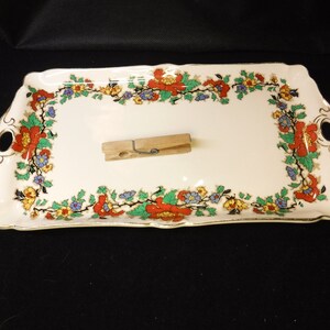 May include: A white rectangular serving tray with a gold trim and a floral design in shades of red, green, yellow, and blue. The tray has two handles on the sides.